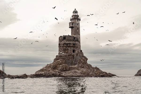 Fototapeta An old crumbling, very beautiful and high lighthouse, on a rock in the open sea, around which gulls and other birds circle in flocks. The grandeur of the past, the incredibly beautiful landscape