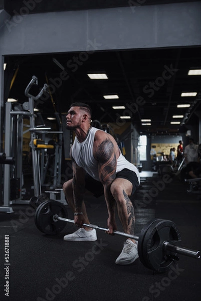 Obraz Vertical full length shot of a strong muscular tattooed male athlete working out at fumctional training gym, lifting barbell