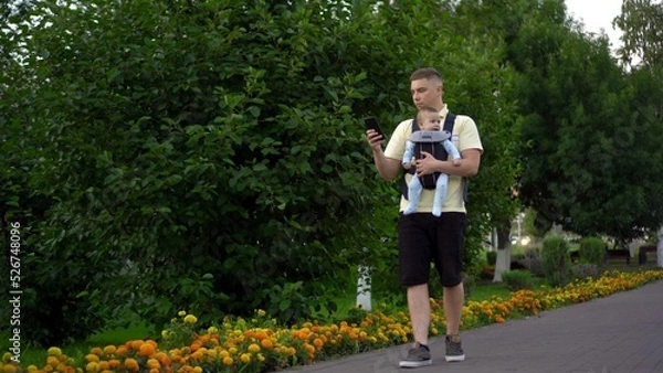 Fototapeta A young dad walks with a child sitting in a kangaroo backpack. A single father walks through the park with a phone in his hands.