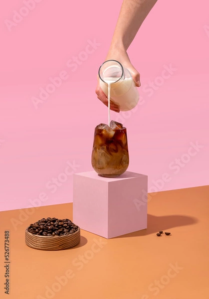Fototapeta Ice coffee in a tall glass with cream poured over and coffee beans. A woman's hand pours cream into a glass of coffee.