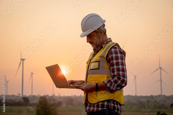 Fototapeta Portrait of engineer wearing yellow vest and white helmet using a computer laptop on site at wind turbines field or farm, Sustainable energy concept