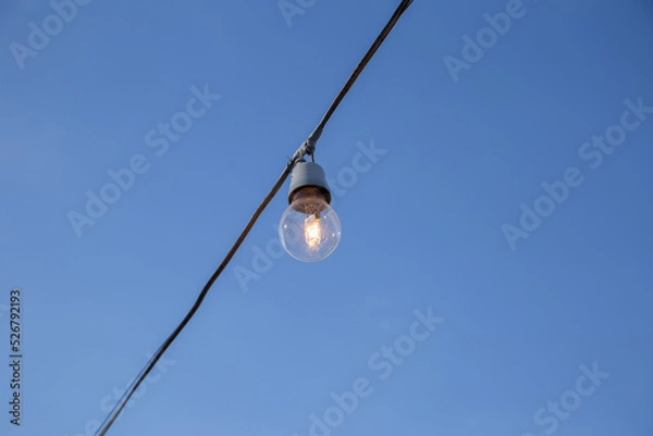 Fototapeta Uma lâmpada conectada a um fio elétrico com céu azul ao fundo.