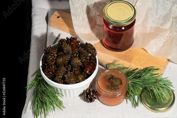 Fototapeta On a black background on a light fabric stands delicious caramel jam and syrup from young small pine cones in a glass jar and a bowl with branches and needles