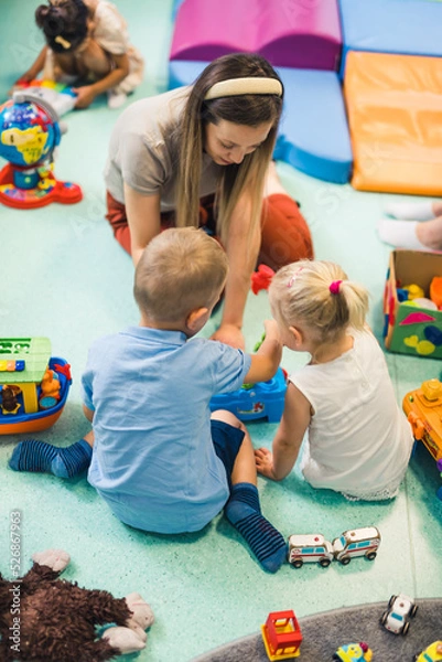 Fototapeta Learning through play at the nursery school. Caucasian toddlers and their teacher playing with colorful plastic playhouses, building blocks, cars and boats. Imagination, creativity, fine motor and