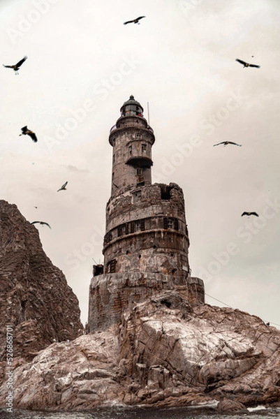 Fototapeta An old crumbling, very beautiful and high lighthouse, on a rock in the open sea, around which gulls and other birds circle in flocks. The grandeur of the past, the incredibly beautiful landscape