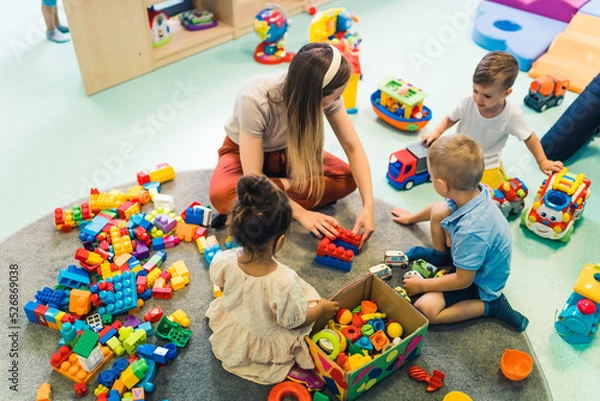 Fototapeta Playtime at nursery school. Toddlers with their teacher sitting on the floor and playing with building blocks, colorful cars and other toys. High quality photo