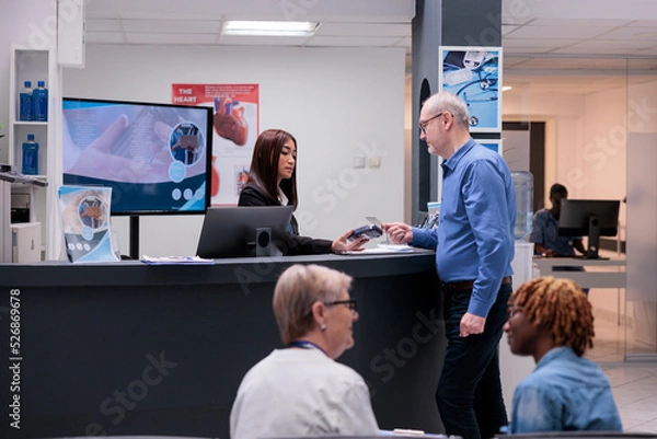 Fototapeta Old man paying consultation with credit card, making electronic payment transaction at hospital reception desk. Having medical checkup appointment at health center, registration counter.