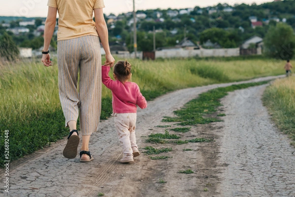 Fototapeta Back view of a mother and daughter walking happy at sunset, holding their hands. Parent, child. Happy family, childhood. Summer nature. Mother nature.