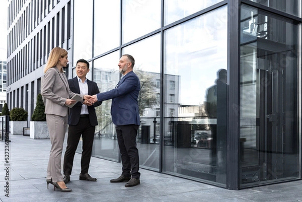 Fototapeta Meeting of three colleagues from outside the office building, experienced and mature IT specialists, greeting and shaking hands, business persons in business suits, diverse group of people