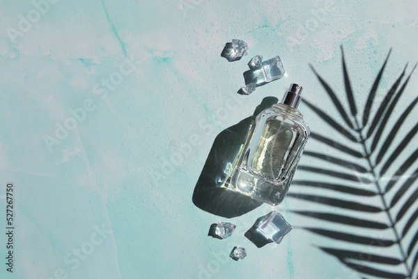 Fototapeta Glass bottle of perfume with shade of palm leaves, ice cubes, water drops on the light blue background. Smell of summer. Fresh  cooling perfumery.