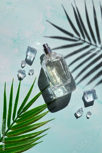 Fototapeta Glass bottle of perfume with shade of palm leaves, ice cubes, water drops on the light blue background. Smell of summer. Fresh  cooling perfumery.