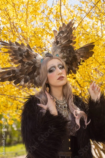 Fototapeta Portrait of a fantastic elf in decorative jewelry in a headdress with feathers on a background of yellow flowers
