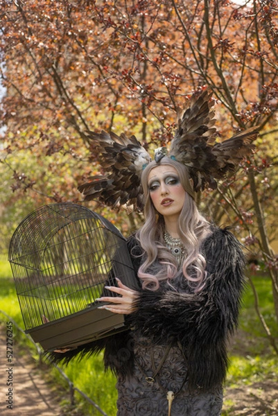 Obraz A girl in a gloomy image with a beautiful make-up, a crown of feathers and a cage in her hands