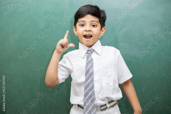 Fototapeta Smart little Indian schoolboy having an idea expression with wide open mouth finally found a solution, blank chalkboard. Education concept.