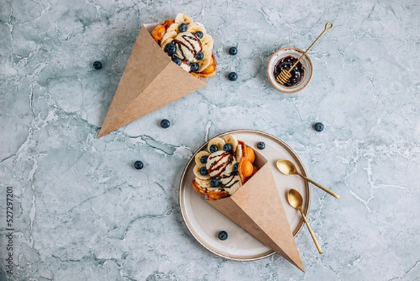Fototapeta Bubble waffle with ice cream, banana and blueberry in paper cup over gray marble background