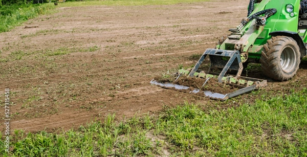 Fototapeta Land work by the territory improvement. Small tractor with a ground leveler for moving soil, turf. A green mini skid steer loader clear the construction site. Machine for agriculture work. Copy space