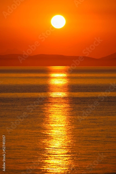 Fototapeta Reflection of the sun rising from behind the mountains on the horizon on the surface of sea water.