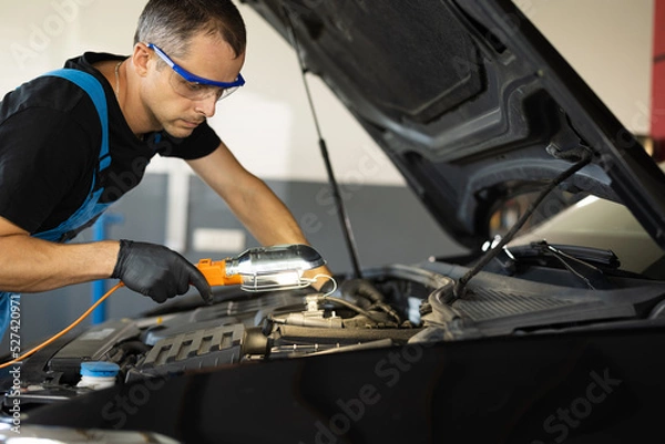 Fototapeta Caucasian Mechanic in Blue Overalls is Working on a Car in a Car Service. Repairman in Safety Glasses is Working on an Usual Car Maintenance. He Hangs Led Lamp. Modern Workshop