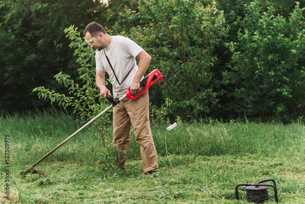 Fototapeta A bearded man is working in the garden. A worker mows the grass with an electric trimmer. Gardener taking care of the lawn