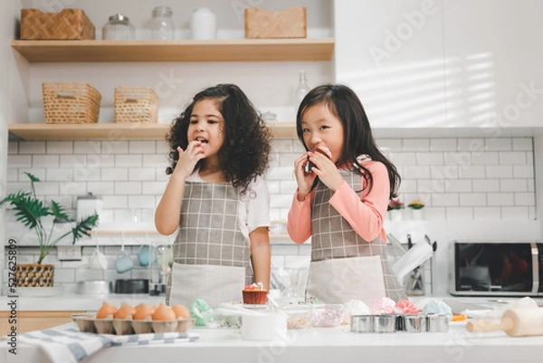 Fototapeta Children enjoy cooking in the kitchen. Happy Asian kids decorating the cake, bake cookies in the kitchen. Two cute little diversity girls play and learn trough the experience. Education concept.