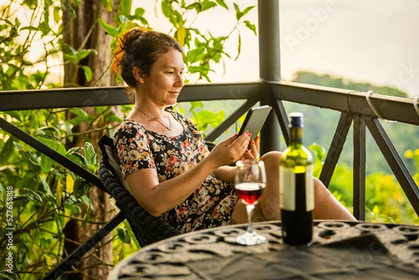 Fototapeta Smiling woman sits on beautiful Costa Rican patio reading an ebook with a glass and bottle of wine