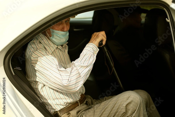 Obraz An old man in a taxi. A man in a medical mask is sitting in a car. Outside view of the pensioner sitting in the car.