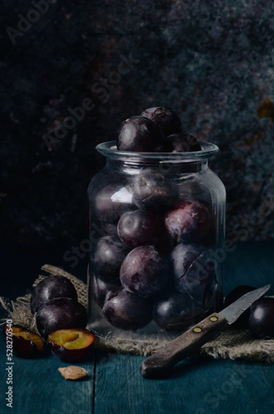Fototapeta A glass jar with dark red plums stands on a blue wooden table, next to it are several plums, one cut and a small old knife