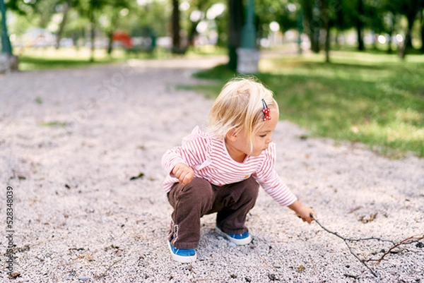 Fototapeta Little girl squats on the gravel with a branch. High quality photo