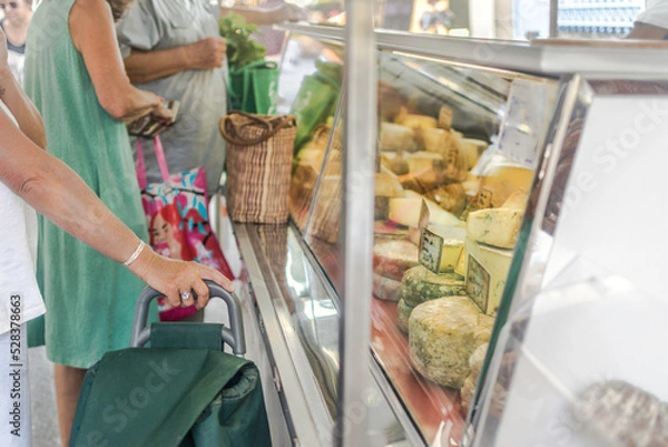 Fototapeta Shoppers in front of French cheese stall at open air farmers' market, outdoors. customer with shopping cart. Buying and choosing a selection of gourmet cheeses.