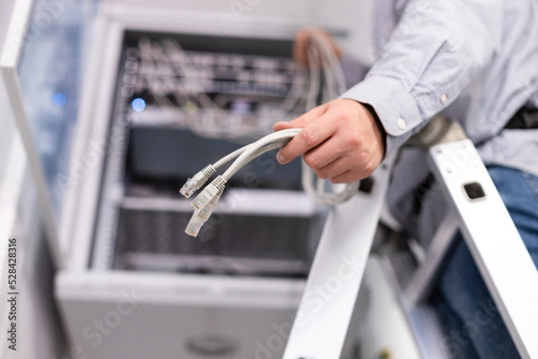 Fototapeta Male worker configures local network by holding cables and plugs to connect to server.