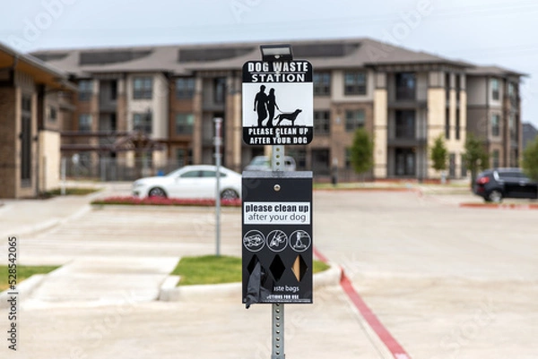 Fototapeta Dog waster station sign with plastic bags for waste. Clean after your dog while walking in residential complex, waste station with baggies with building and car parking on background.