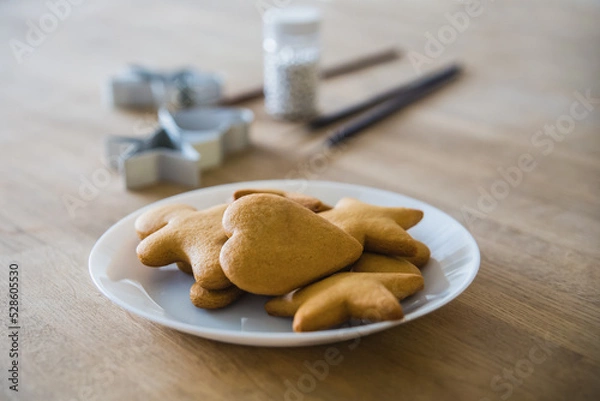Fototapeta A white plate of traditional ginger biscuits stands on a wooden table. Brushes for drawing on gingerbread, forms for gingerbread in the form of hearts and stars.