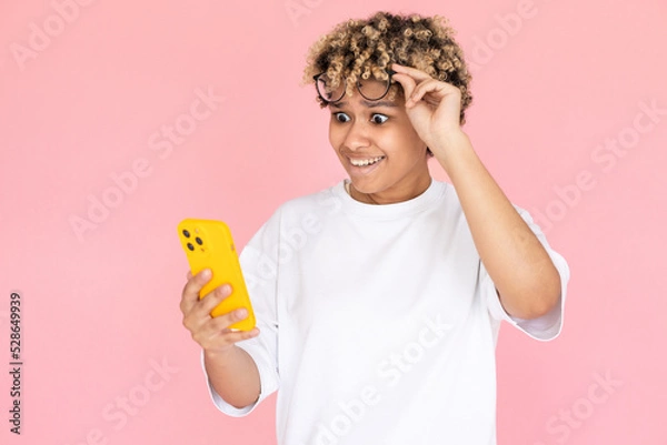 Fototapeta Portrait of amazed African American woman. Female model in glasses with curly hair looking at mobile phone with shock. Portrait, studio shot, emotion concept