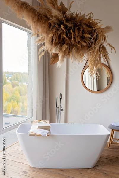 Fototapeta Comfortable bright bathroom with a boho-chic interior design, a free-standing white bath against the background of a panoramic window. hanging decor from dry grass. Soft selective focus.