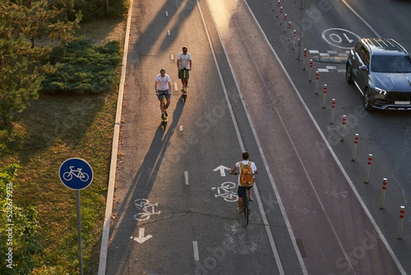 Fototapeta Top aerial view of cyclist and two mans are riding on kick scooters on cycling road.