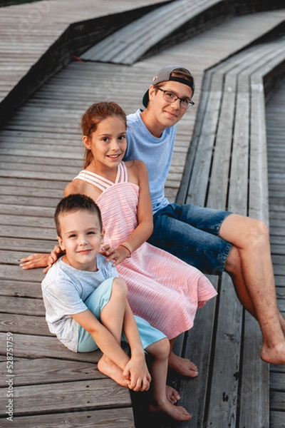 Fototapeta three caucasoid teenagers from the same family of different ages sister with two brothers on wooden benches together side by side smiling at sunset in summer with bare feet