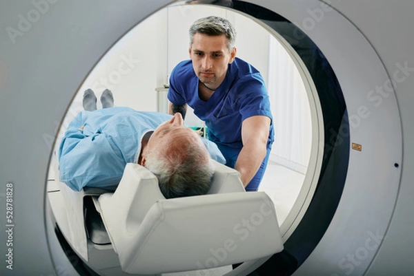 Fototapeta Senior man going into CT scanner. CT scan technologist overlooking patient in Computed Tomography scanner during preparation for procedure