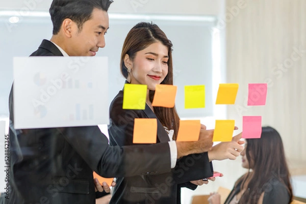 Fototapeta Asian young beautiful professional successful female businesswoman in formal suit standing smiling holding hand pointing presenting information data on sticky note on glass board to colleague