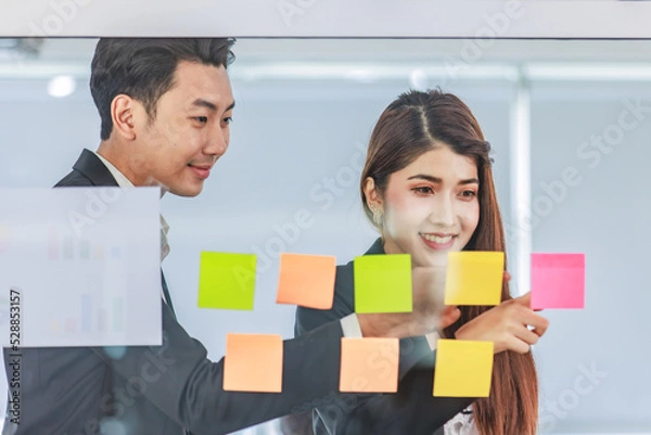 Fototapeta Asian young beautiful professional successful female businesswoman in formal suit standing smiling holding hand pointing presenting information data on sticky note on glass board to colleague