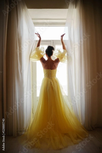 Fototapeta A woman's silhouette in a golden luxurious dress against the background of a window holds a curtain with her hands. Elegant lady in a yellow long silk dress with bare back, back view.