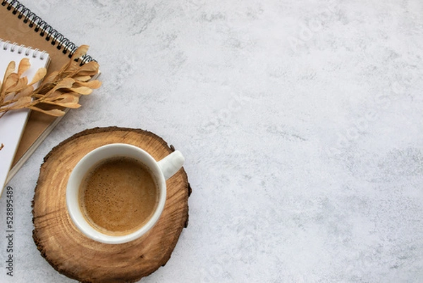 Fototapeta Cup of cappuccino coffee on tree cut wooden cup coaster, spiral notebook and kraft paper sketchbook on office table background. Top view, flat lay, copy space. Stationery supply

