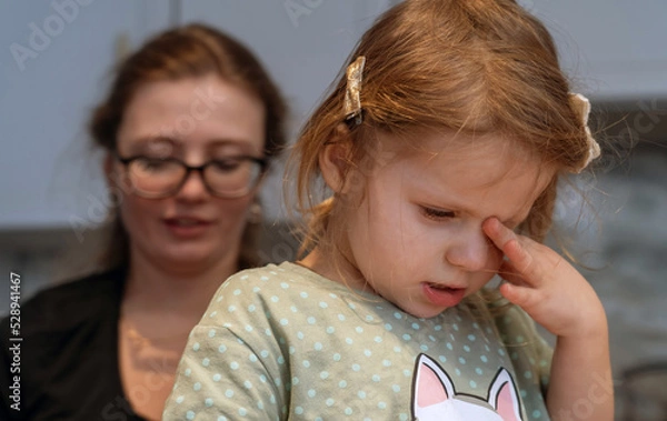 Fototapeta Sleepy, moody little girl stands in kitchen and rubs eyes while her mom is on phone on social networks. Mother plays on phone but does not play with her daughter