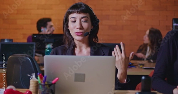 Obraz Confident young woman speaking with headset in front of computer screen at office call center. Female employee working at customer help desk talking