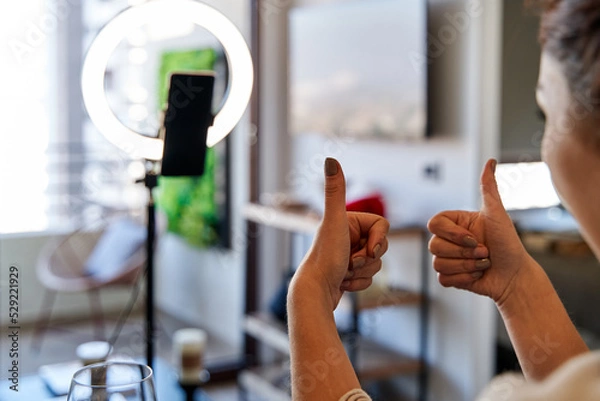 Obraz young female influencer sitting in the dining room in front of her smartphone and a ring of light, showing her followers her hands in approval. social media concept