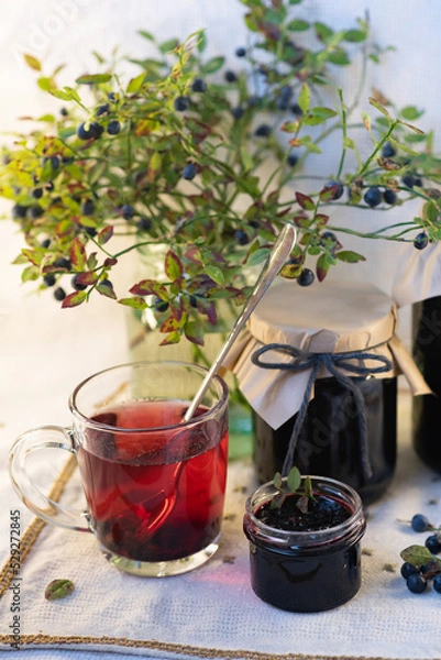Fototapeta On a light background, on a beautiful fabric napkin, there are several packaged glass jars with blueberry jam, and in front there is a fresh tasty blueberry with green leaves on the branches and a swe