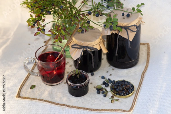 Fototapeta On a light background, on a beautiful fabric napkin, there are several packaged glass jars with blueberry jam, and in front there is a fresh tasty blueberry with green leaves on the branches and a swe