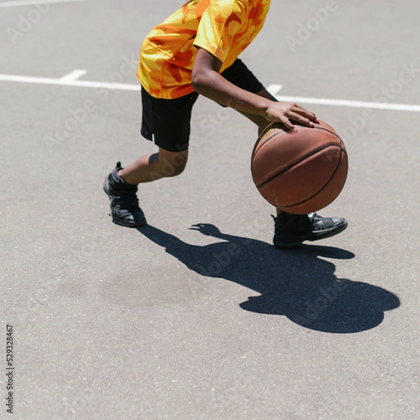 Fototapeta Unrecognizable dark skinned kid with a yellow sports uniform plays basketball on a sidewalk on a urban street. There are no trademarks in the shot.