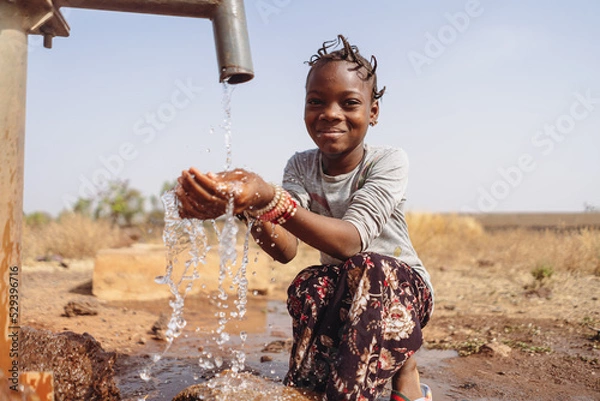 Obraz Thirsty African girl with funny braids, sitting in the middle of a puddle, happy to collect the abundant water that flows from the village fountain; concept of water scarcity in developing countries