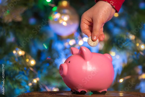 Fototapeta Children's hands put a coin in a pink piggy bank on a New Year's bokeh background. The concept of investment in the future of the child, savings, financial planning, fulfillment of desires