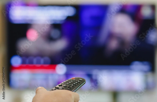 Fototapeta Close-up of hand with the remote control television and presses the button. Television remote control changes channels thumb on the blue TV screen.